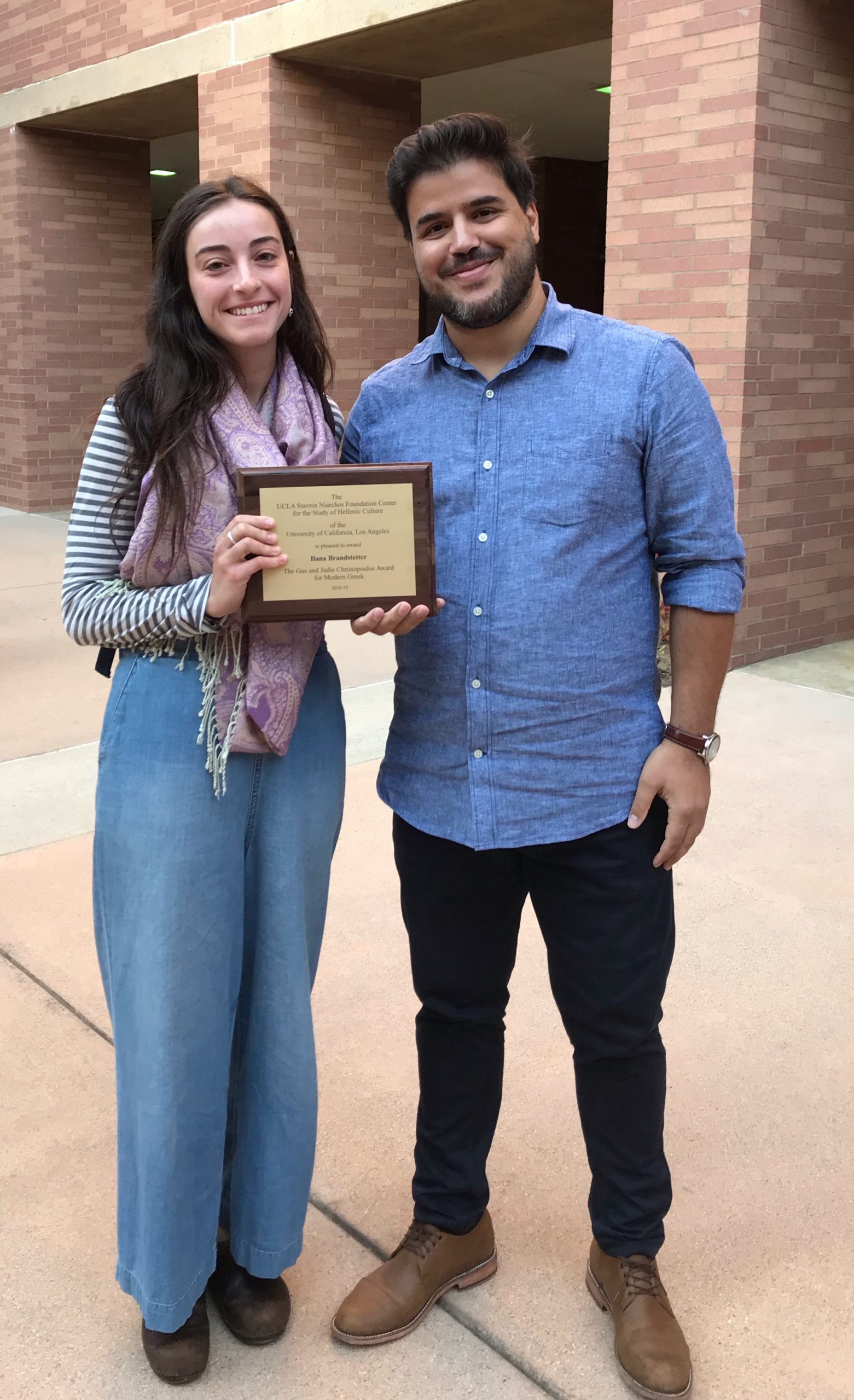 Image of Ilana Brandstetter with Dr. Simos Zenios. Ilana Brandstetter is holding up a plaque. The building in the back is a brick building on the UCLA Campus.