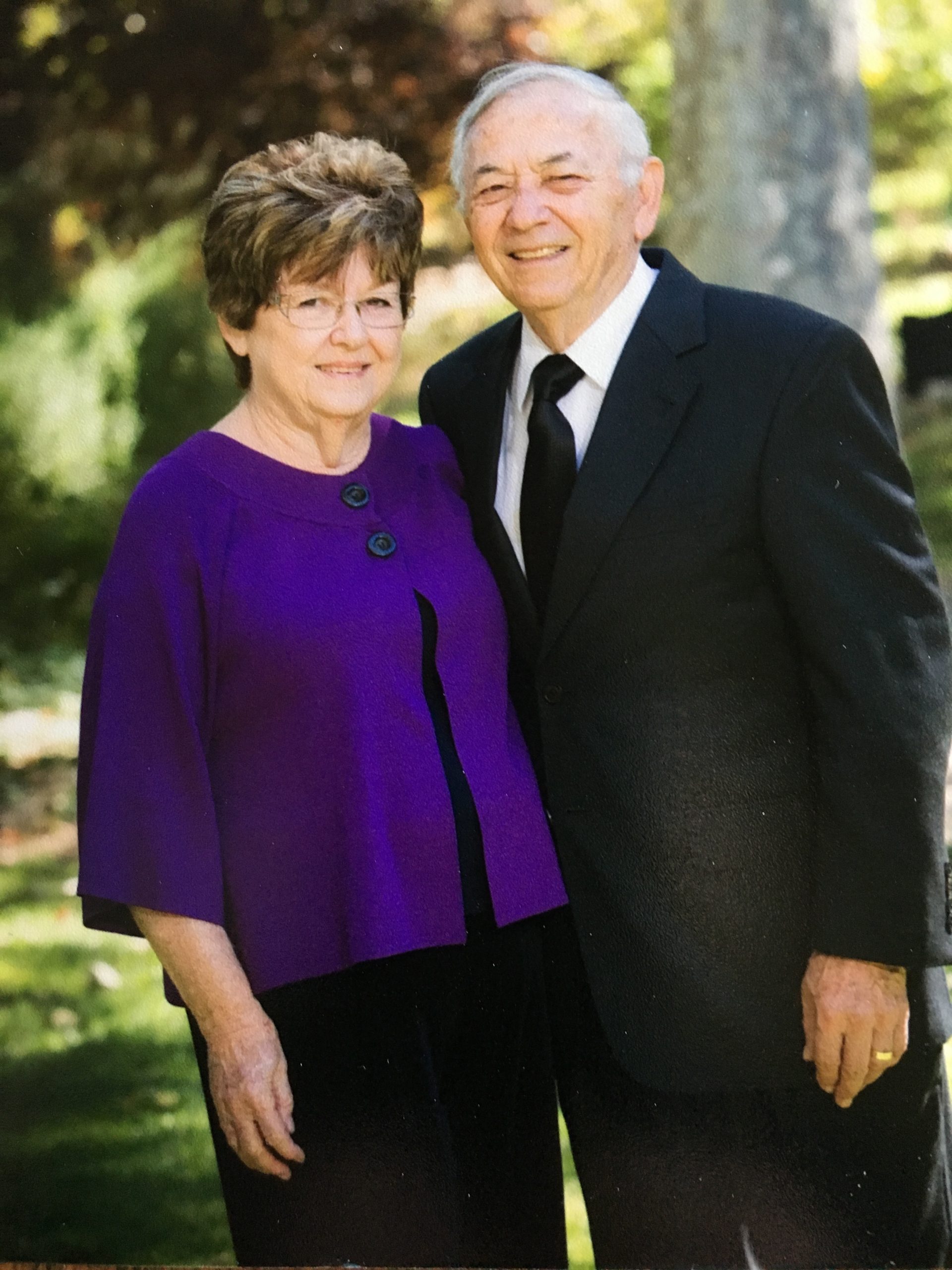 Photo of James and Carolyn Kolokotrones. Carolyn Kolokotrones is wearing a purple cardigan and a black dress. James Kolokotrones is wearing a black suit with a white dress shirt and black tie. They are outside standing in front of a tree.