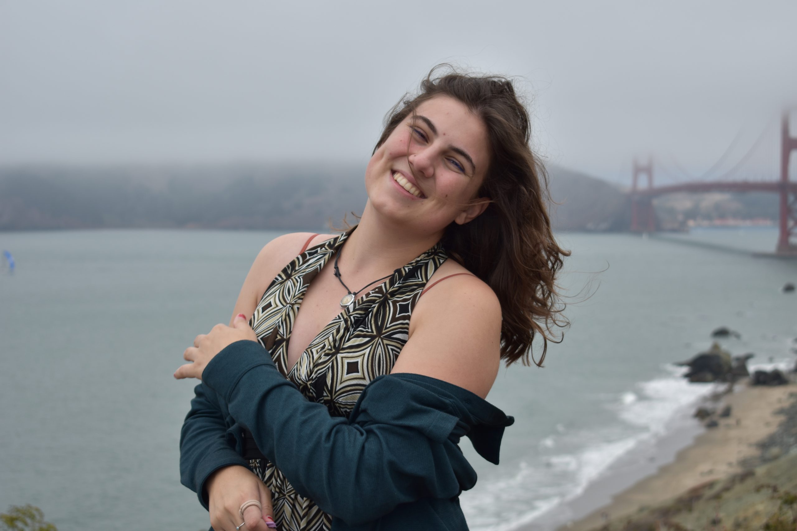 Myrsini San Marchi standing on the coast near the Golden Gate Bridge, wearing a necklace, a brown and tan dress, and a blue jacket, with foggy skies and hills in the background.