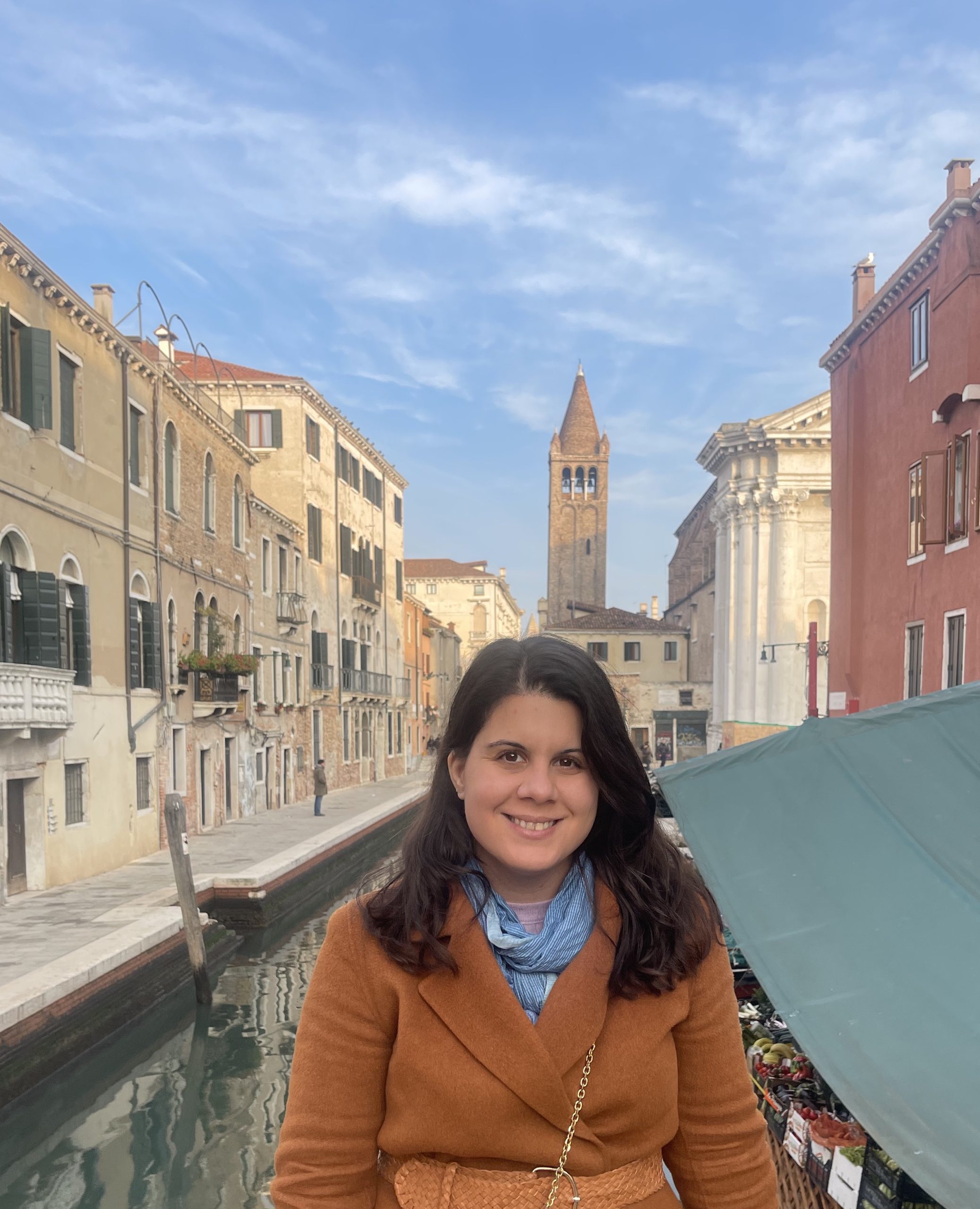 Image of Leah Marangos. She is wearing a blue scarf and a camel toned coat. She is sitting on a boat in a canal in a city.