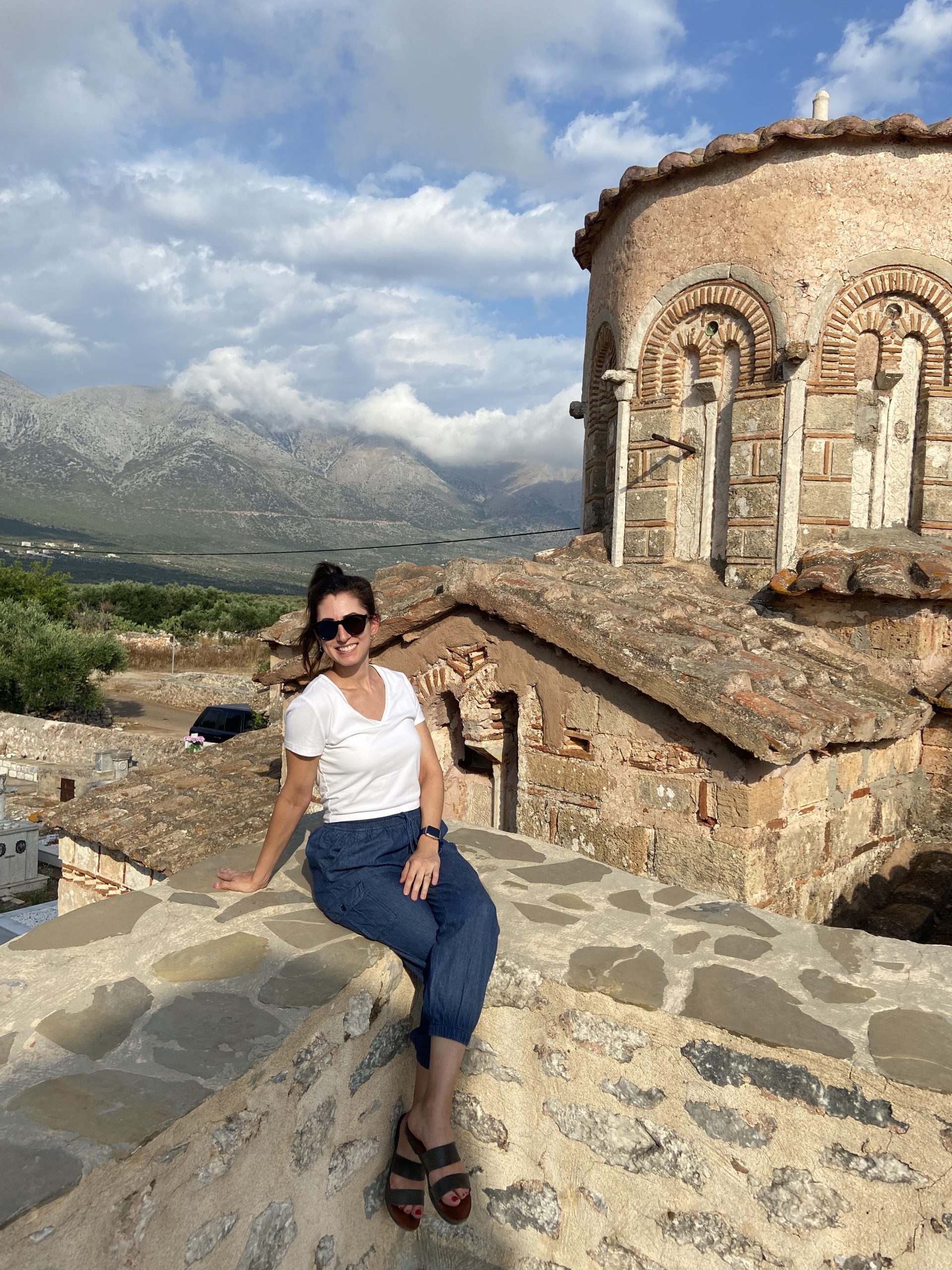 Image of Christine Muron sitting outside a stone balcony. There is a Greek chapel near her. Hills and clouds are in the background.