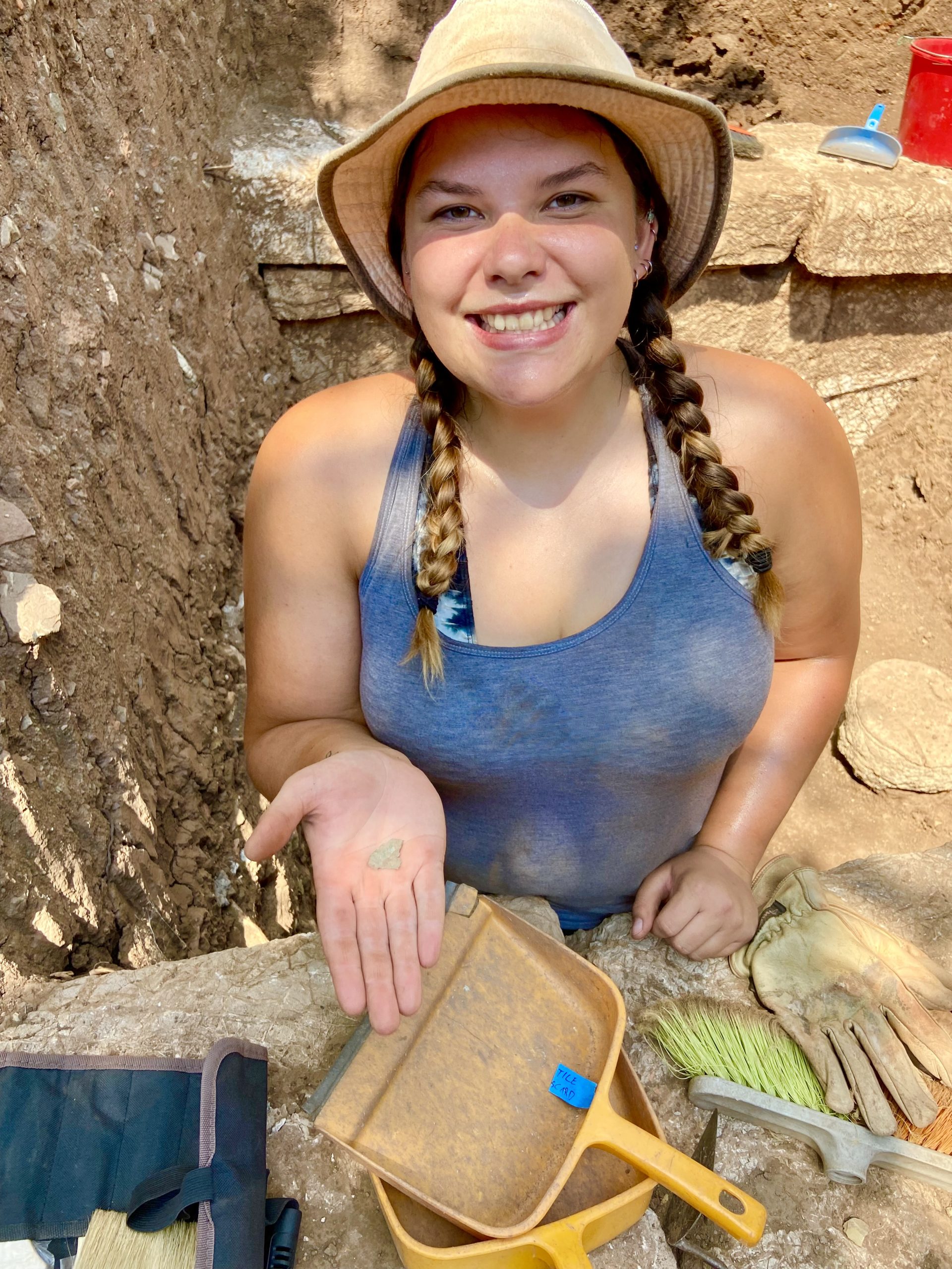 Katrina Kuxhausen-Derose at an archaeological dig site, wearing a tan hat and gray shirt, with yellow dust pans, a yellow brush, and gloves laid out in front of her.