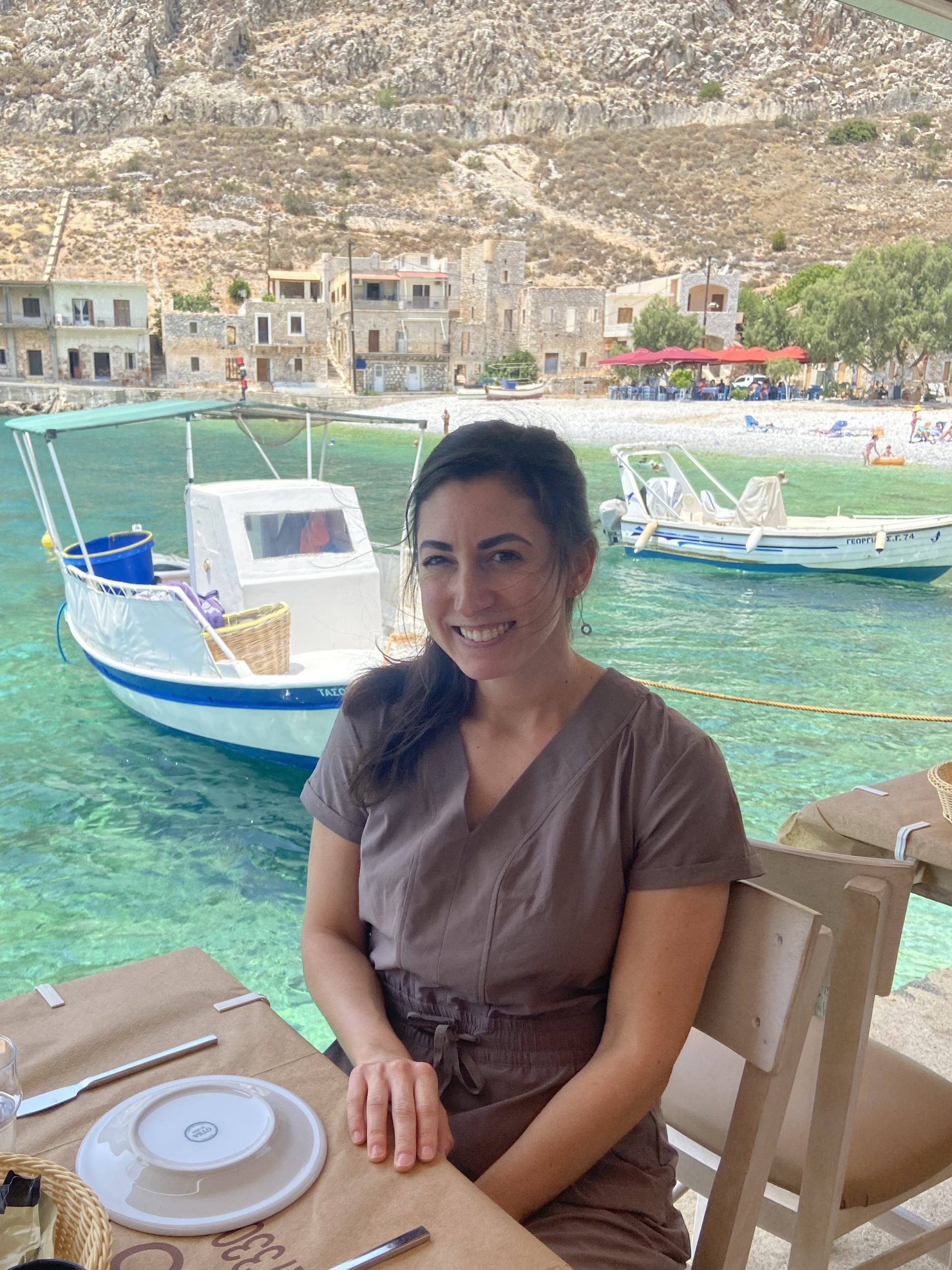 Photo of Christine Muron wearing a brown dress with the coastline of Mani, Greece, in the background. Stone buildings line the shore, and boats are visible on the blue-green water.