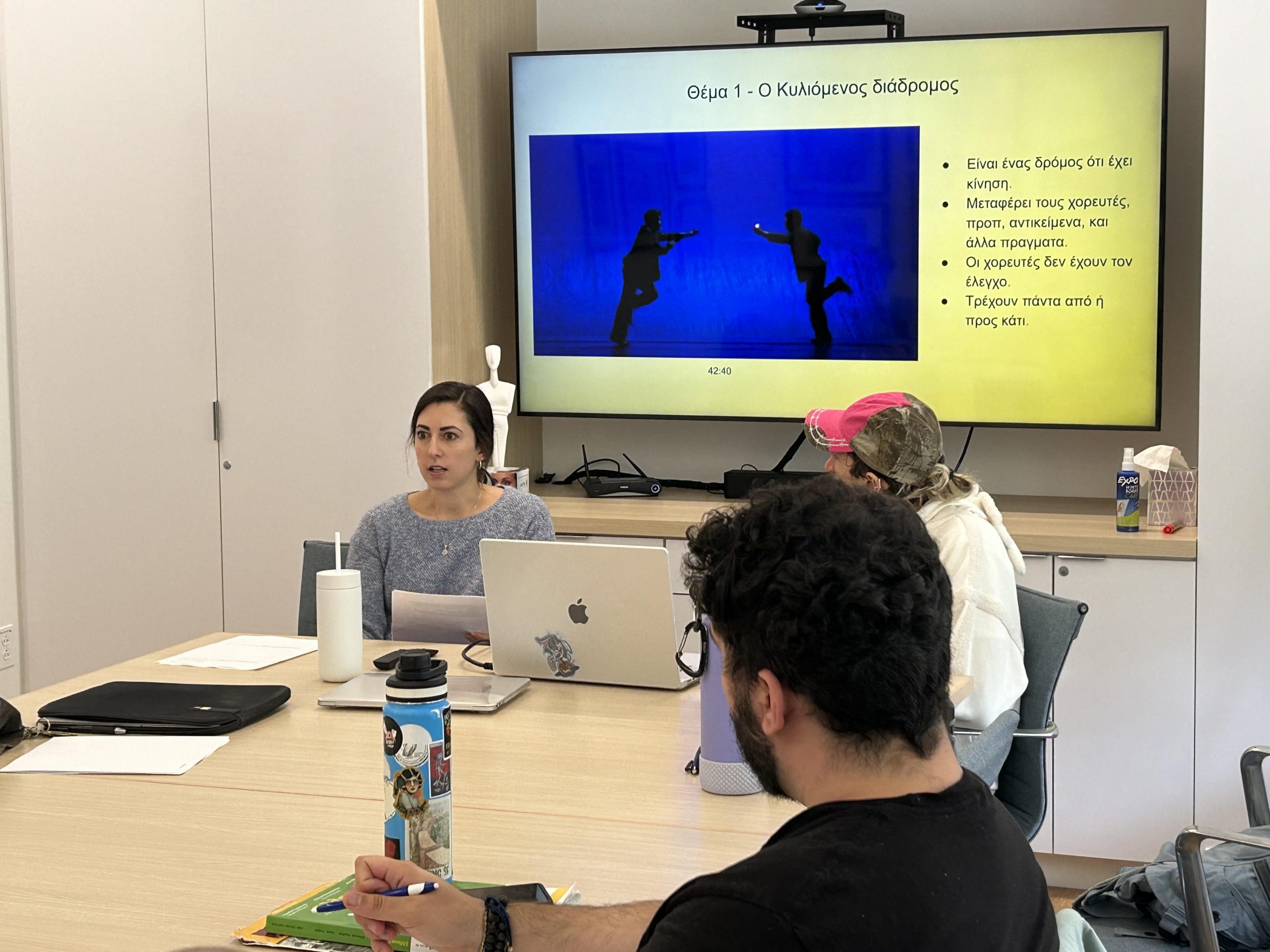 Image of Christine Muron and other students presenting on Dimitris Papaioannou. There is a television screen with a PowerPoint on the screen.