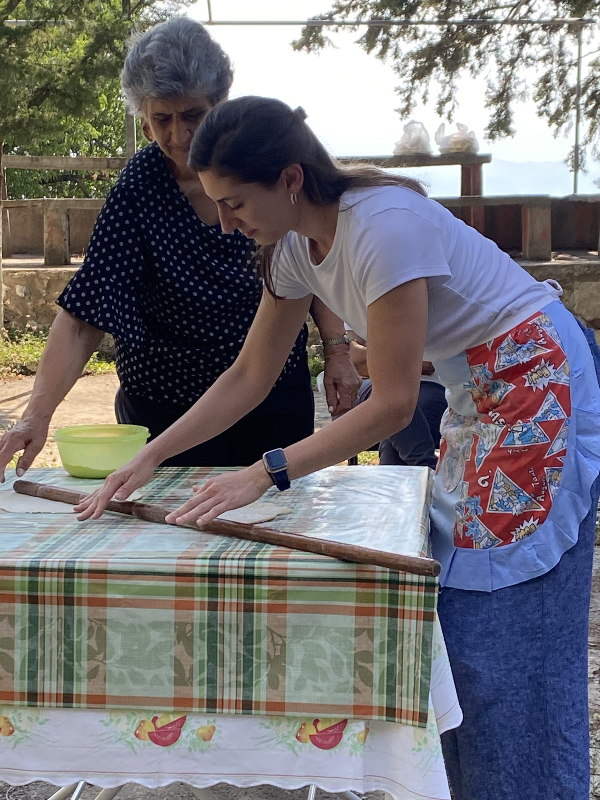 Image of Christine Muron and a woman from Geraki. They are baking for the community. The student is holding a rolling pit and it wearing an apron. The table has a plaid table cloth and they are outside amongst trees.