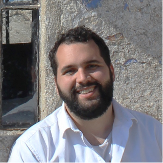 A photo of Luis Rodriguez-Perez. He is wearing a white long sleeved shirt. He is standing in front of a stucco structure with a window partially in view.